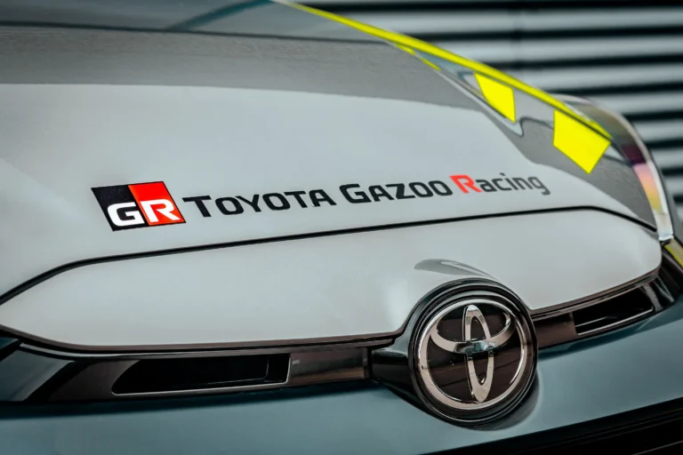 Nahaufnahme des Toyota Gazoo Racing GR-Emblems auf matt-grauer Motorhaube; Carbon-Optik und fluoreszierende Livery betonen den turboaufgeladenen Hot-Hatch-Charme.